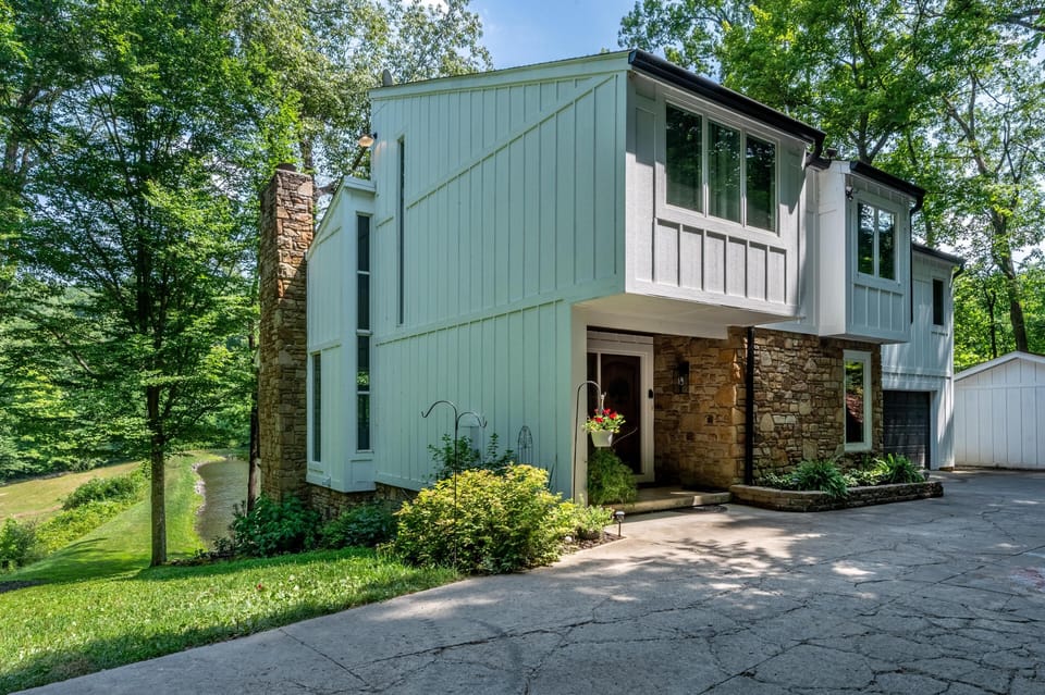 White vertical siding and stone exterior walls flank large windows, with a concrete driveway and lush greenery.