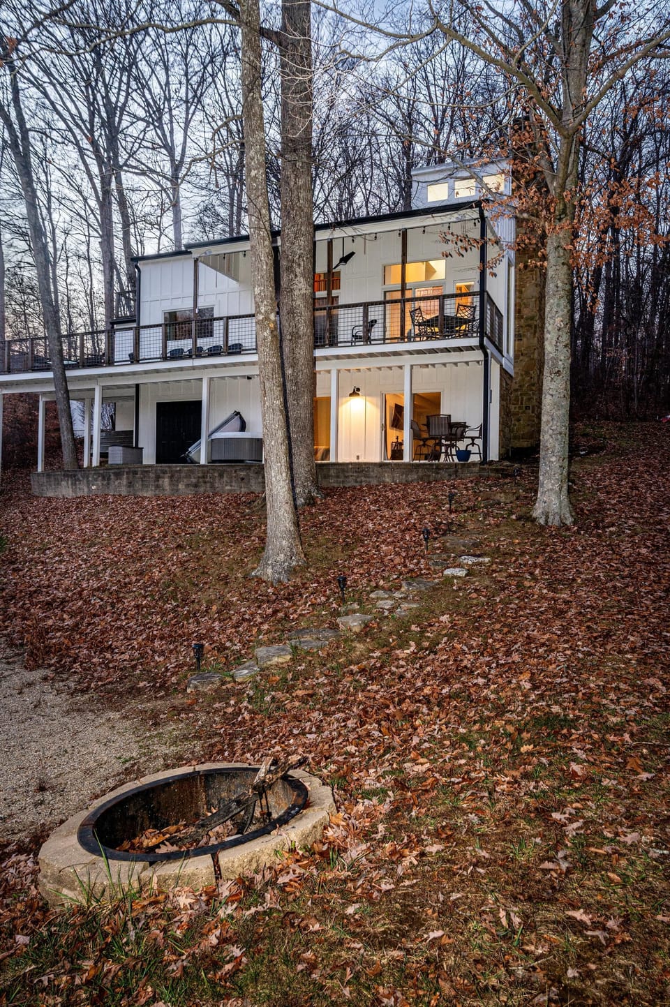 Exterior view of white vertical siding house with stone chimney, covered balconies, and a stone fire pit in leaf-covered yard.