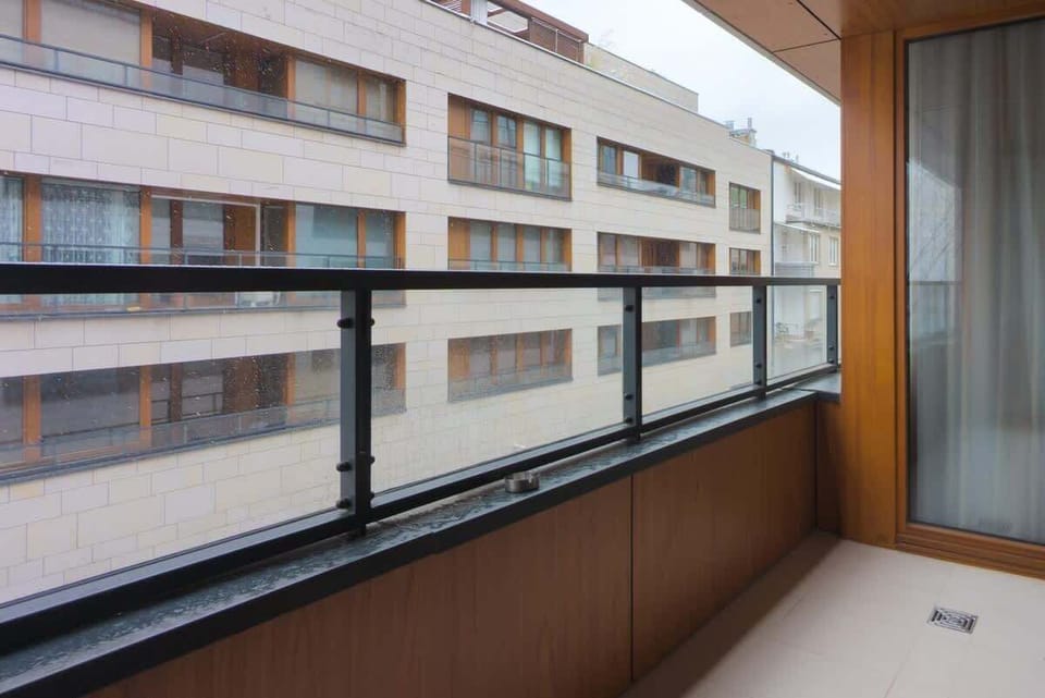 A balcony with wooden paneling and a protective glass railing, offering an urban view of adjacent buildings.

