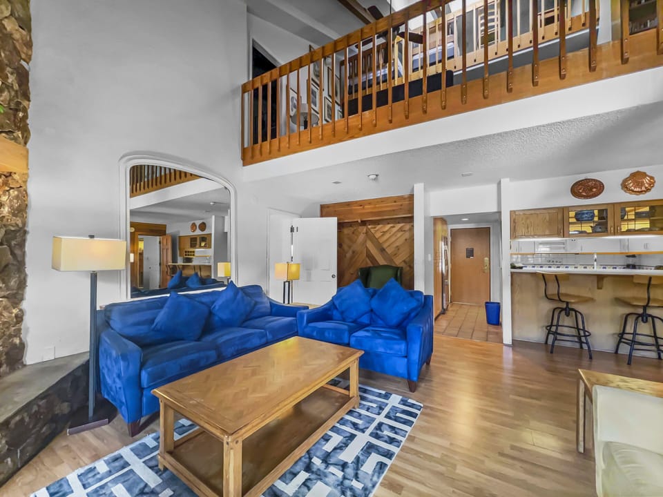 A living room with blue sofas, wooden coffee table, and rug. A loft with a wooden railing is shown above, and a kitchen area with bar stools is visible in the background.