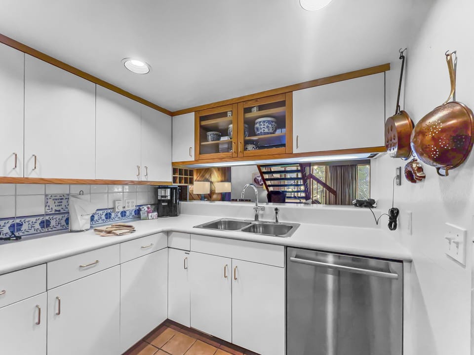 A kitchen with white cabinets, stainless steel sink and dishwasher, copper pots hanging on the wall, and blue and white dishware in a glass-front cabinet.