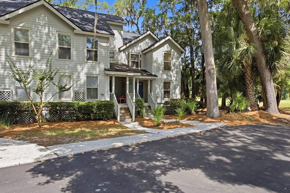 Spanish moss drapes elegantly over towering oaks, casting dancing shadows across this quiet coastal home. Tropical greenery and a crisp white porch create a peaceful entry point for your next getaway under the bright blue sky.