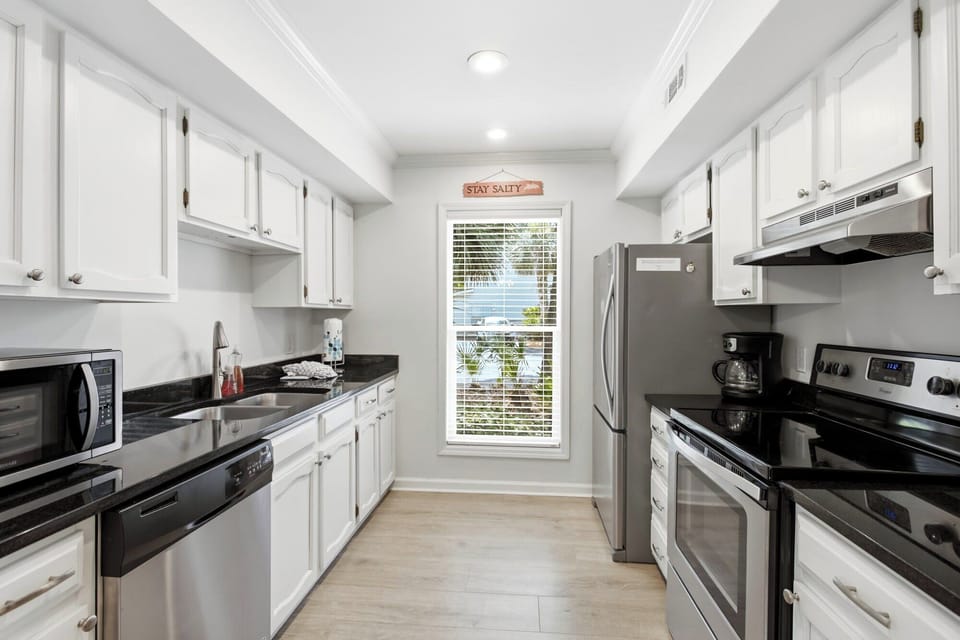 Polished granite counters and crisp white cabinetry create a sleek, airy kitchen for your morning brew. Natural luminescence pours through the tall window, highlighting the clean lines and modern stainless steel appliances.