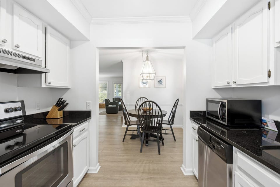 Crisp white cabinetry and sleek granite counters frame a passage to the sun-drenched dining area. Soft overhead glow and warm wood floors create a seamless, inviting flow for your morning coffee.