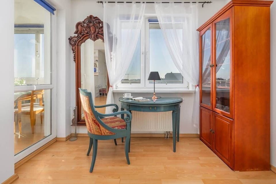 A bright corner of the apartment with a wooden floor, a classic-style desk and chair, sheer white curtains, and an ornate mirror, complemented by a tall wooden wardrobe on the right.
