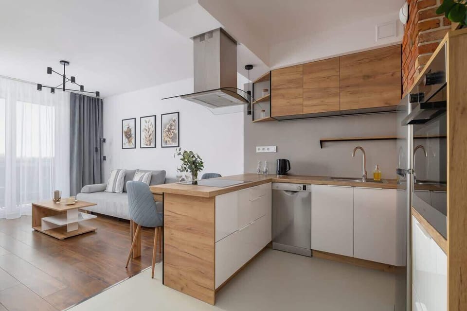 A compact open-plan kitchen with light wooden cabinets, grey countertops, and a small dining area with minimalist decor.

