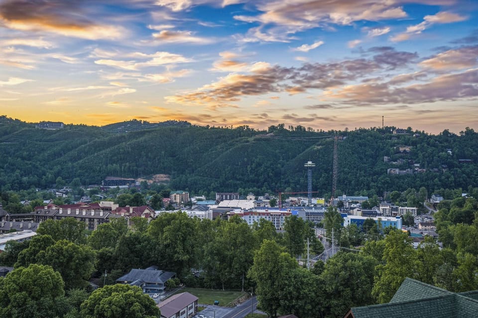 Panoramic hillside neighborhood with lush greenery and distant city buildings framed by a stunning sunset sky painted in vibrant amber and blue hues.