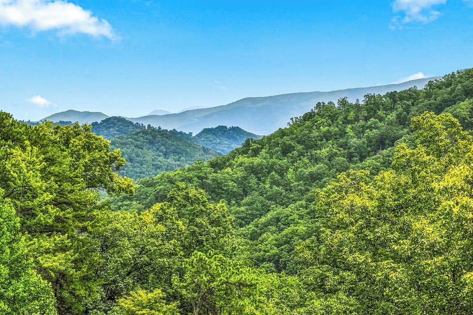 A scenic mountain landscape captured from the property, with rolling forested ridges stretching into the distance beneath a bright blue sky and gentle clouds.