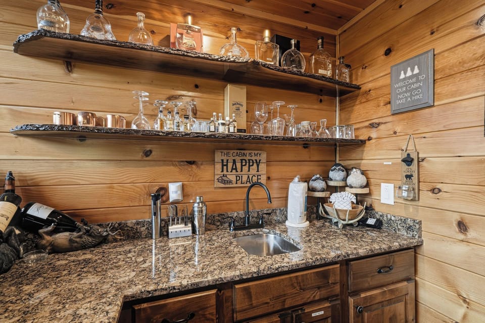 Your cabin retreat features a welcoming wet bar with granite countertops, rustic wooden shelving displaying glassware, and charming cabin-themed décor—perfect for unwinding with your favorite drink.