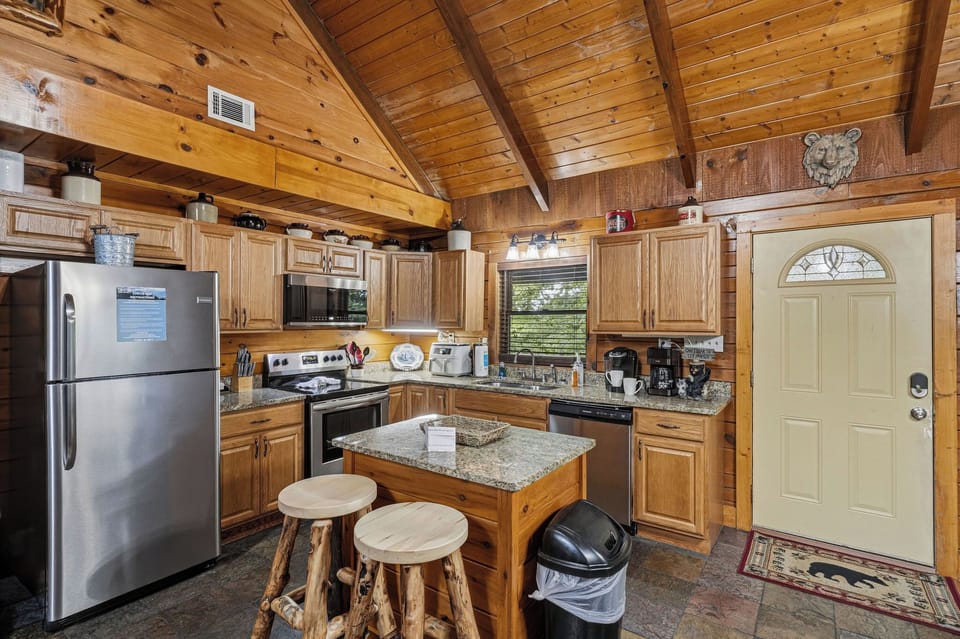 Step into your rustic cabin's fully equipped kitchen, where soaring vaulted ceilings and warm wood paneling create a cozy mountain retreat atmosphere. The granite island with rustic log stools invites you to gather and cook in comfort.