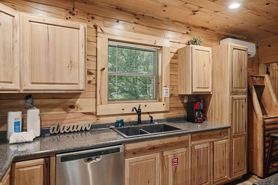 Step into your rustic cabin kitchen with warm pine cabinetry, granite counters, and modern appliances. Natural light streams through the window above the farmhouse sink as greenery peeks in from outside.