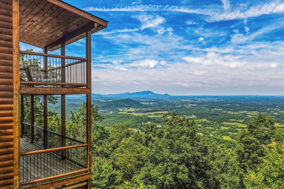 The multi-level balcony structure overlooks expansive valley farmland and distant mountain ridges beneath a dramatic blue sky, offering a scenic backdrop to the property's elevated forest setting.