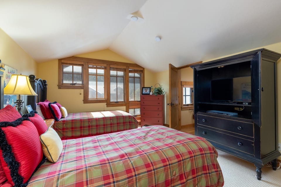 A bedroom with two plaid-patterned twin beds, a large black TV cabinet, a red dresser, and angled windows letting in natural light.