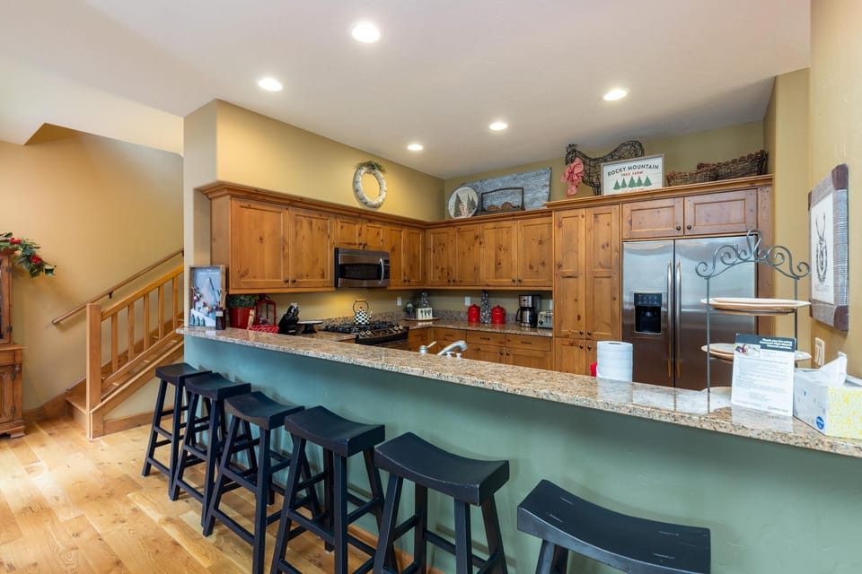A kitchen with wooden cabinets, an oven, microwave, stovetop, and refrigerator. A long counter with a granite top has five black barstools. Decorative items are displayed on the upper cabinets.