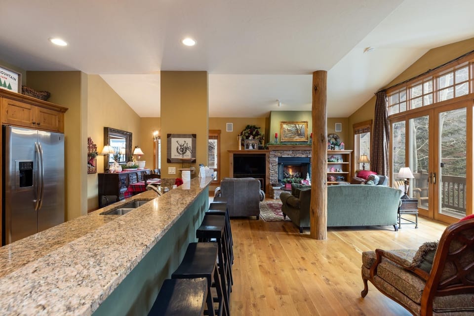 A cozy living room with a stone fireplace, a green sofa, an armchair, and a large window. Adjacent kitchen area features a long granite countertop with barstools and stainless steel appliances.
