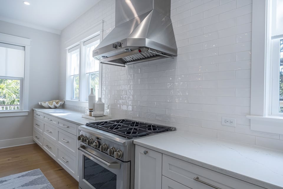 Stainless steel oven and stove with hood above. White cabinets and white tile back splash to the ceiling.