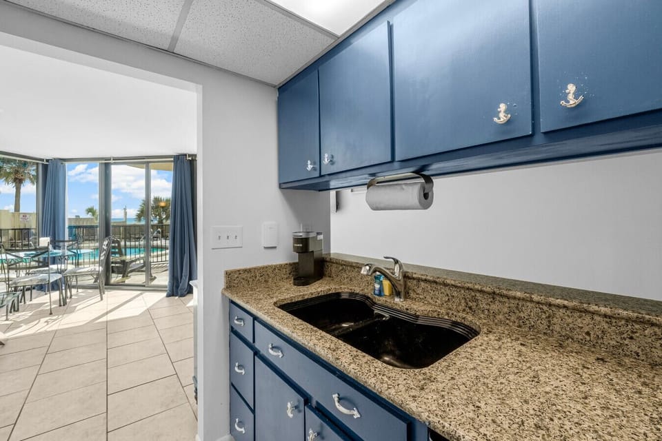 Charming kitchen with granite countertops and navy cabinets opens up to a dining area with stunning poolside and beach views through large glass doors