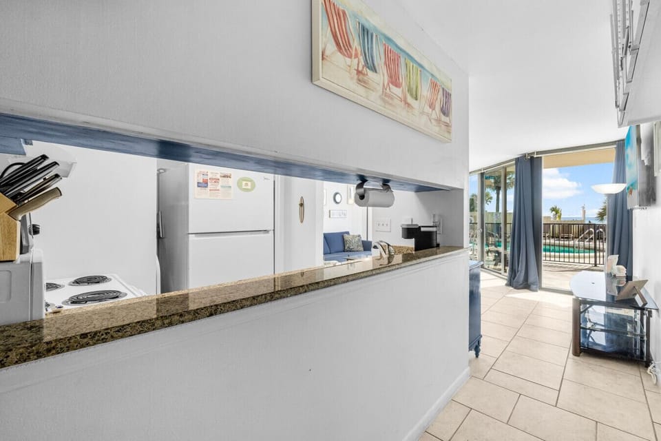 Cozy living space features fully equipped kitchen with granite countertops, bright dining area, and sliding glass doors leading to private patio with poolside and beach views.