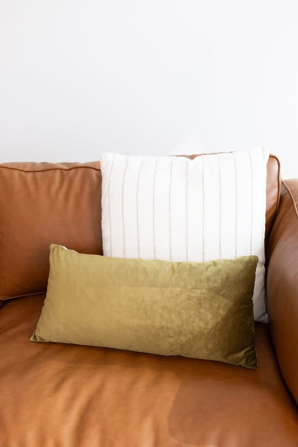 Tan leather sofa styled with a textured white cushion and olive velvet lumbar pillow. Warm tones and soft layering invite you to sink in with a book, a film, or quiet conversation after a day out.