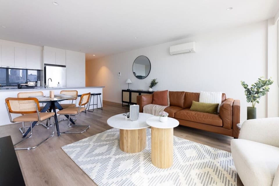 Warm open-plan living and dining area with a tan leather sofa, fluted timber coffee tables, rattan dining chairs, and a white kitchen. A round mirror, indoor plants, and air conditioning complete the comfortable space.  *(216 characters)*