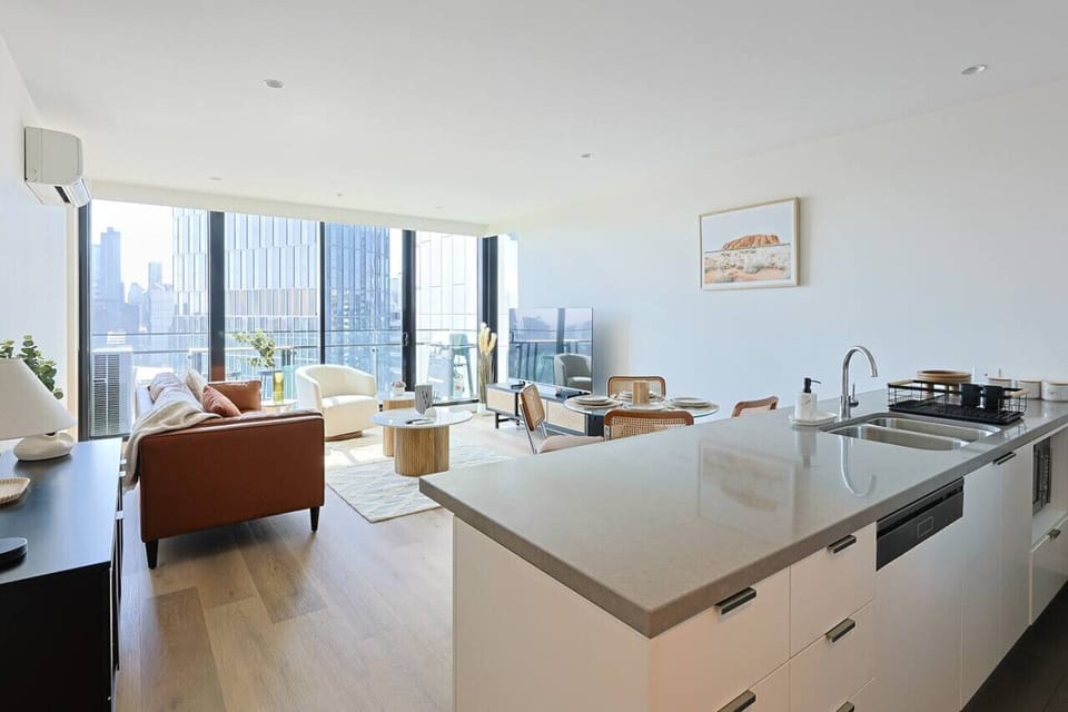 Open-plan kitchen with a stone island bench flowing into a warm living and dining area with tan leather sofa, rattan chairs, and floor-to-ceiling windows framing sweeping city views beyond.  *(189 characters)*