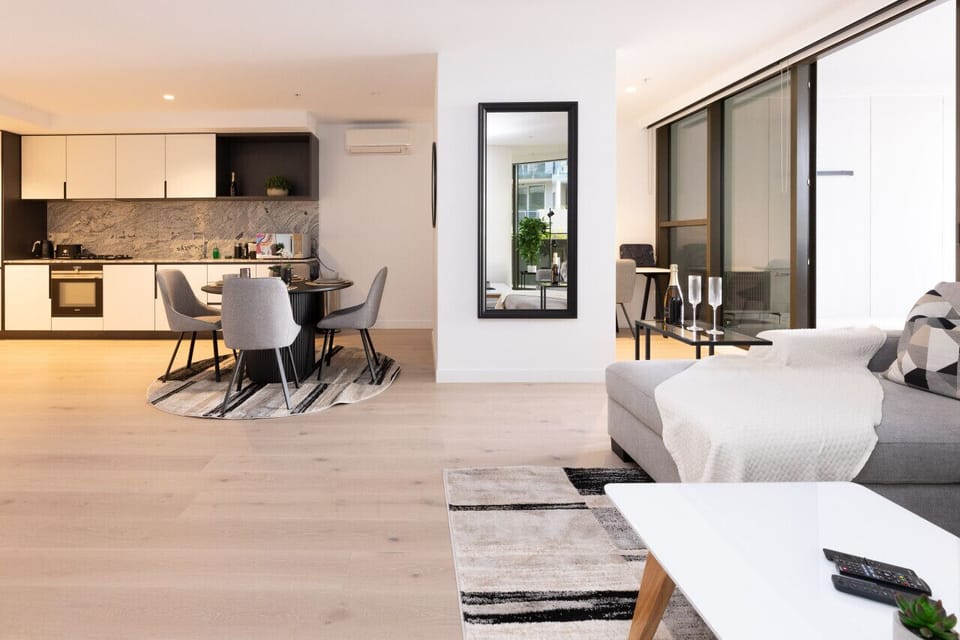 Expansive open-plan layout connecting the living area, round dining table, and fully equipped kitchen with marble-look splashback. Light timber floors, a full-length mirror, and balcony access make the space feel airy and connected.  *(230 characters)*
