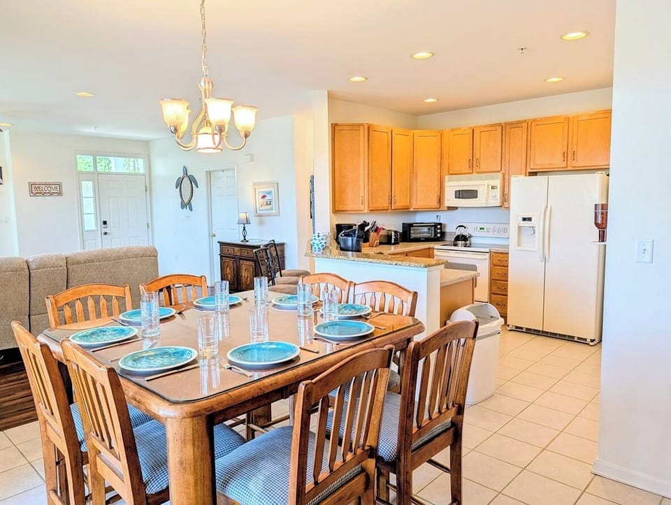 128C October Glory Dining Room to Kitchen View - Open-concept dining space at 128C October Glory in Bear Trap Dunes, Ocean View, DE, showcasing a wooden dining set with turquoise place settings, a classic chandelier, and a seamless view into the adjacent kitchen with light wood cabinets and white appliances.