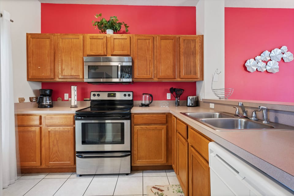Bright and functional, this kitchen features rich wooden cabinetry paired with sleek stainless-steel appliances, all set against a vibrant pink accent wall. Enjoy the spacious countertops, perfect for meal prep or morning coffee.