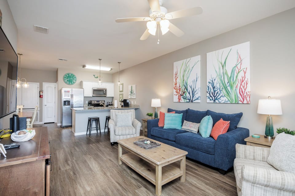 Relax in your bright, coastal-inspired living space with a plush navy sofa, charming sea-themed artwork, and an open kitchen perfect for preparing meals.