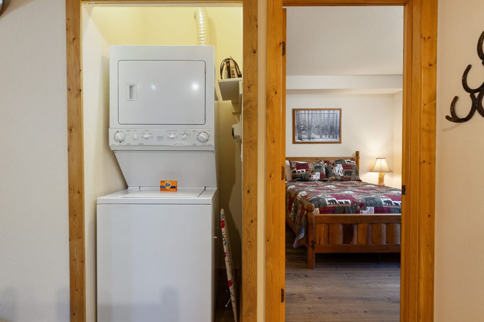 A small laundry area with a stacked washer and dryer is situated between two wooden doorways, leading to a view of a cozy bedroom.