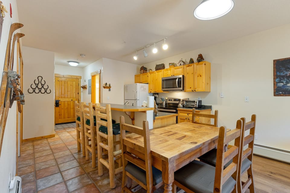 A rustic kitchen and dining area with wooden cabinets and furniture. The kitchen features modern appliances including a refrigerator, microwave, and oven. The room has a tiled floor and overhead lighting.