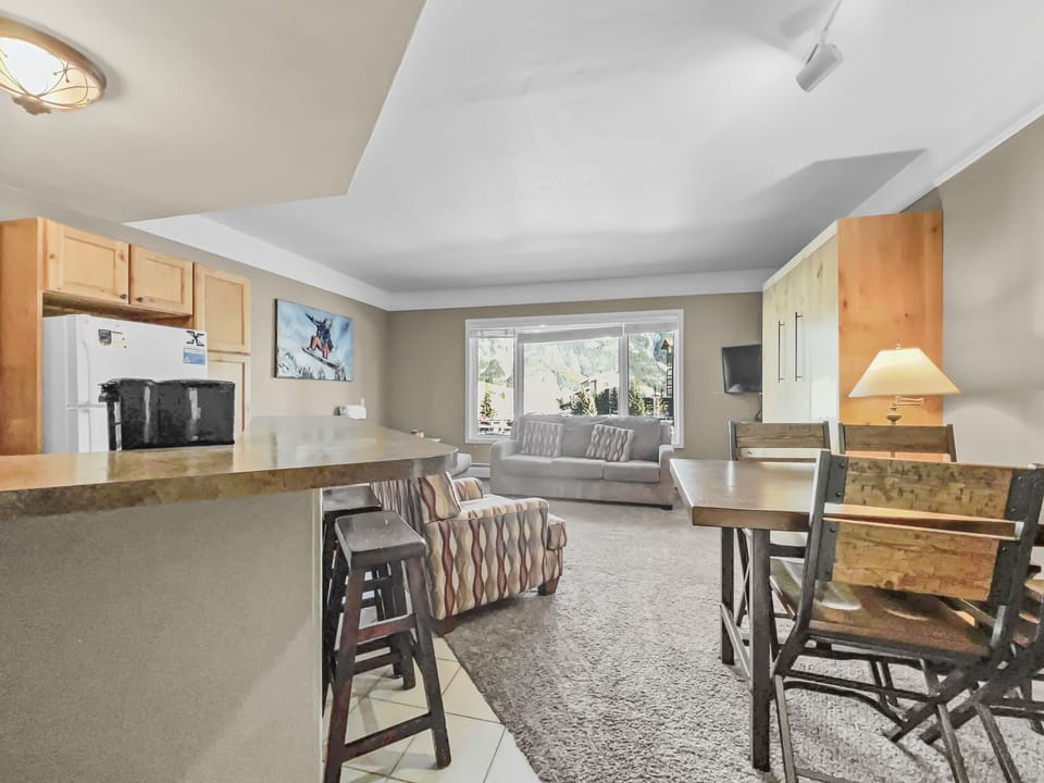 A cozy living area with a kitchen. A countertop with stools, a sofa, armchairs, a dining table with chairs, a window with a scenic view, and a wall-mounted TV are present. Carpeting covers the living area floor.