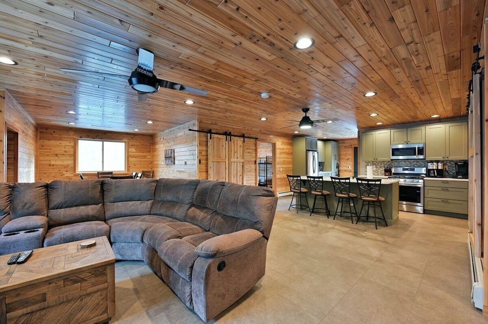 The Taj Woodside has a spacious, modern living area with a large grey sectional sofa, wood-paneled walls and ceiling, and a kitchen island with seating in the background.