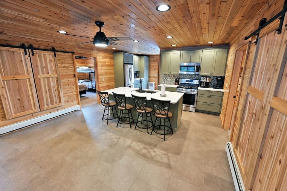 The Taj Woodside has an open-concept cabin interior featuring wood-paneled walls and ceiling, a kitchen with a large island and granite countertops, and sliding barn doors.