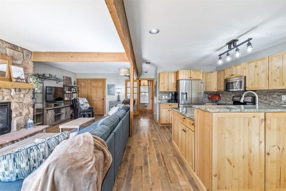 Open-concept living and kitchen area with wooden cabinets, granite countertops, stainless steel appliances, and a stone fireplace. Sofa and TV setup in the living area. Wooden beam ceiling feature.