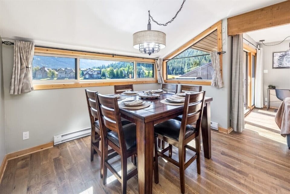 A dining area with a wooden table set for six people, surrounded by windows that offer a scenic view of mountains and houses. The room has wooden floors and a ceiling light fixture.