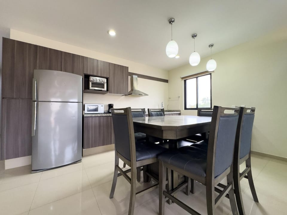 Dining area features a square table with six cushioned chairs beneath three pendant lights by a window.