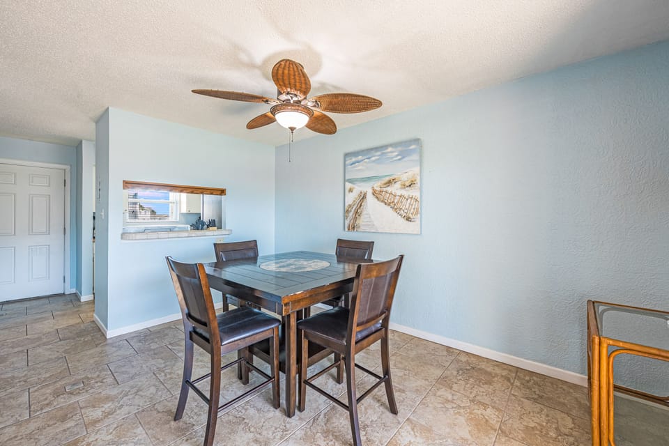 Your coastal dining space features a welcoming table for four, perfect for sharing meals while enjoying the soothing beach artwork and tropical ceiling fan overhead.