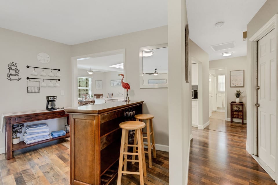 A modern kitchen area with wooden floors, a bar counter with stools, a wall-mounted clock, and a coffee station. There's also a hallway leading to adjacent rooms.