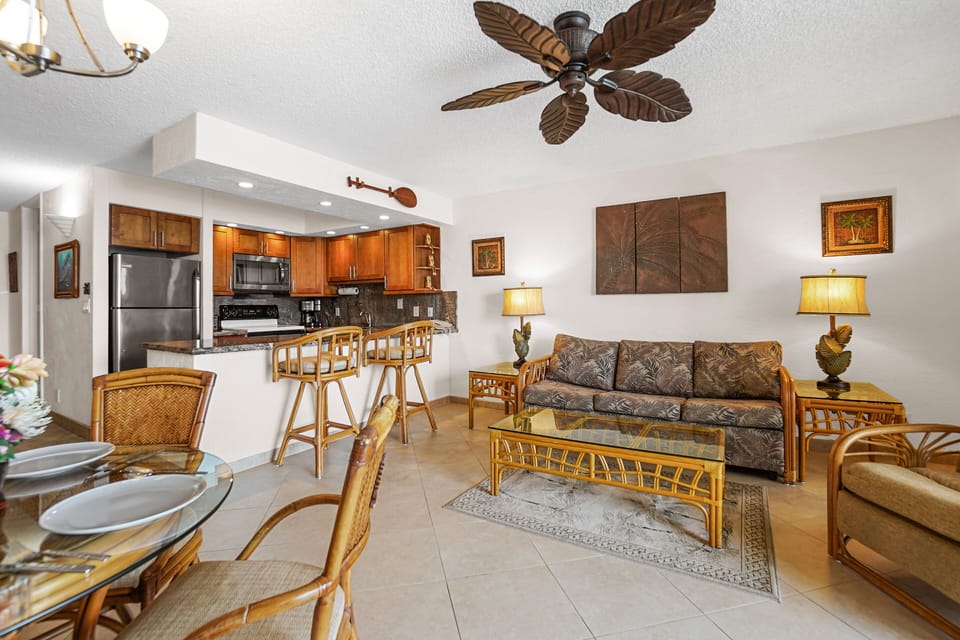 A bright and airy living room featuring rattan furniture, tropical-inspired decor, and a seamless view of the modern kitchen. A perfect island getaway ambiance.