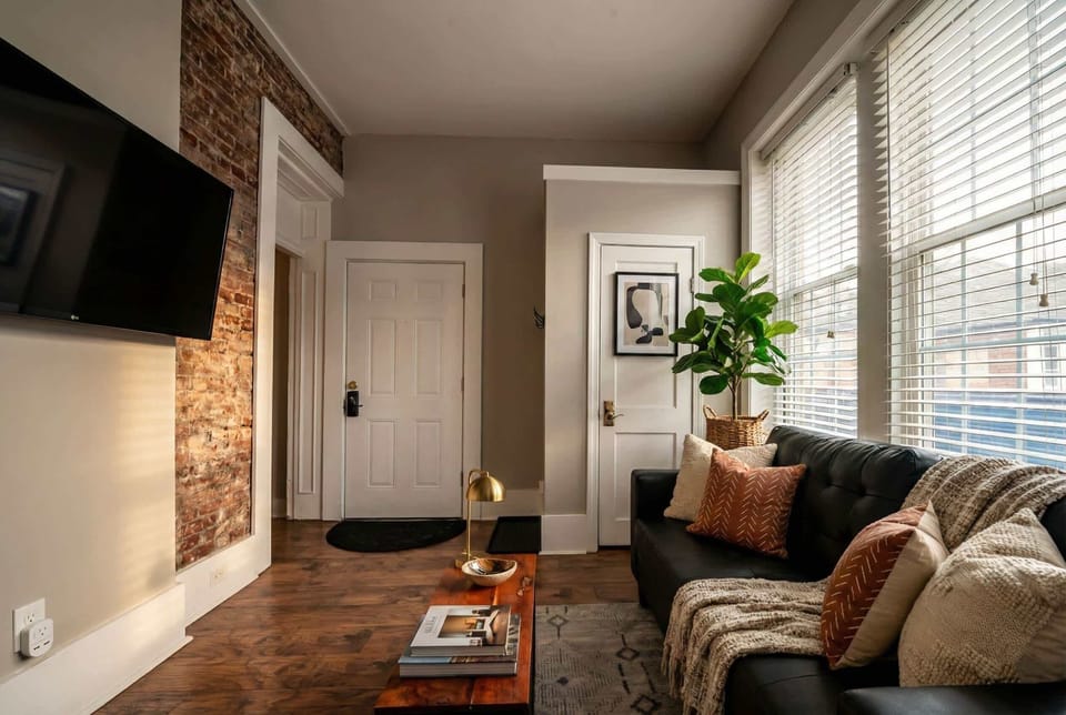 Living room with exposed brick accent wall, sofa, wall-mounted TV, and front entry.