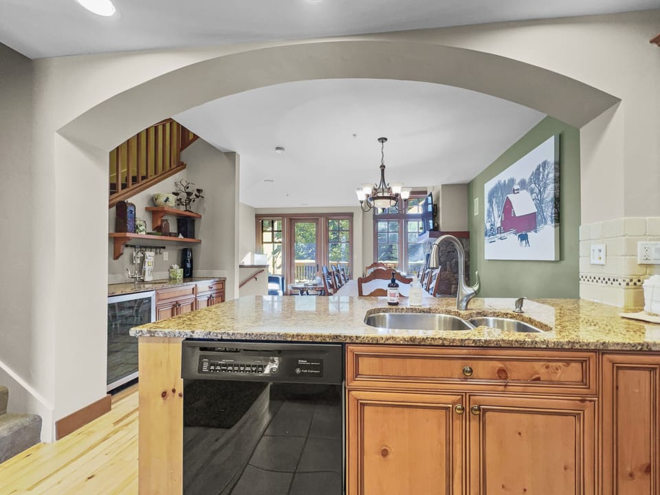 A kitchen with a granite countertop, wooden cabinets, a sink, and a dishwasher. An archway leads to a dining area with a table, chairs, and a painting of a barn on the wall.