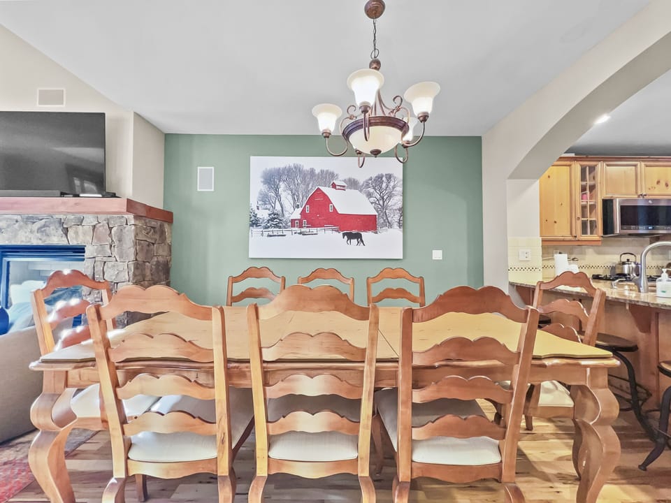 A dining room with a wooden table and chairs, a chandelier, a green accent wall with a painting of a barn, and a fireplace on the left. A kitchen area is visible in the background on the right.