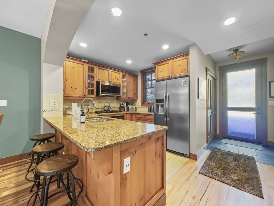 A modern kitchen with wood cabinets, a granite island, stainless steel appliances, and wooden stools at a breakfast bar. A door at the end leads outside, with a mat placed on a stone tile floor.