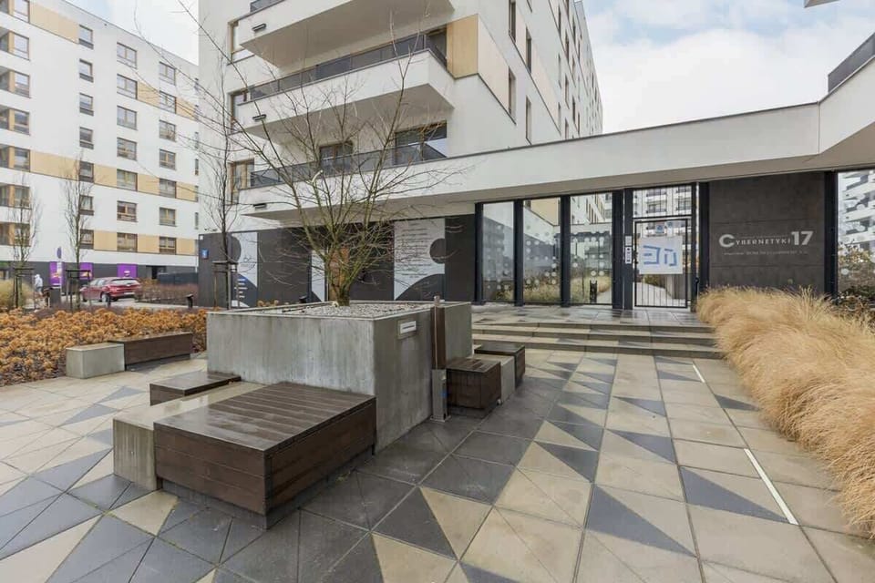 A communal outdoor seating area with wooden benches and tables, surrounded by landscaped gardens and walkways.
