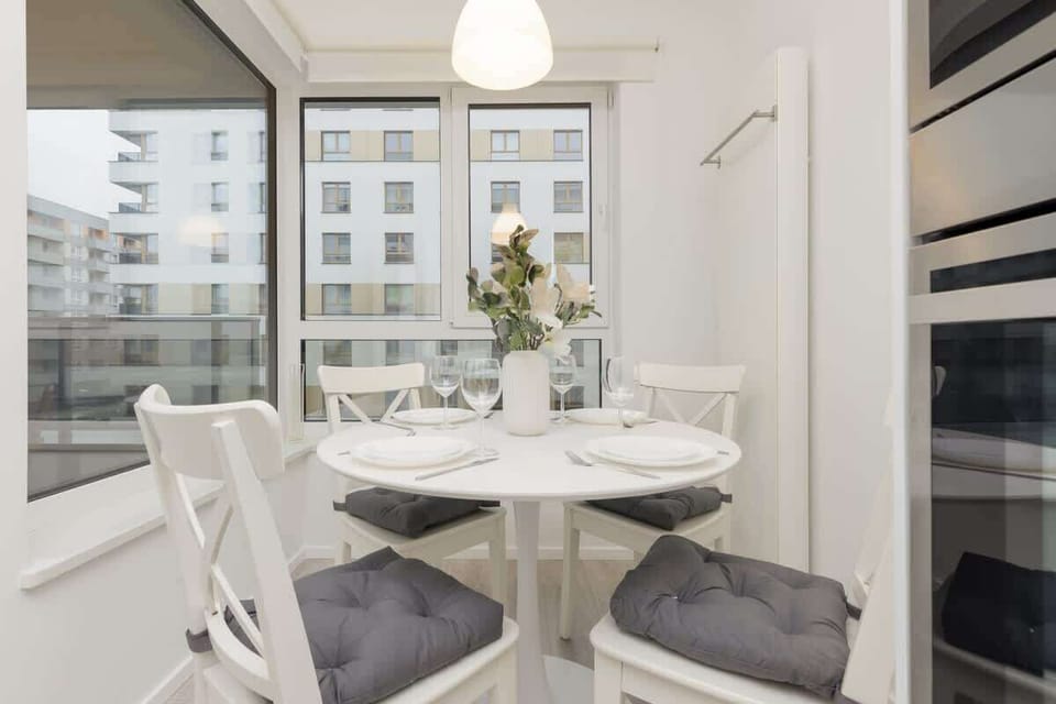 A dining area with a round white table, grey cushioned chairs, and large windows allowing natural light, perfect for meals with a view.
