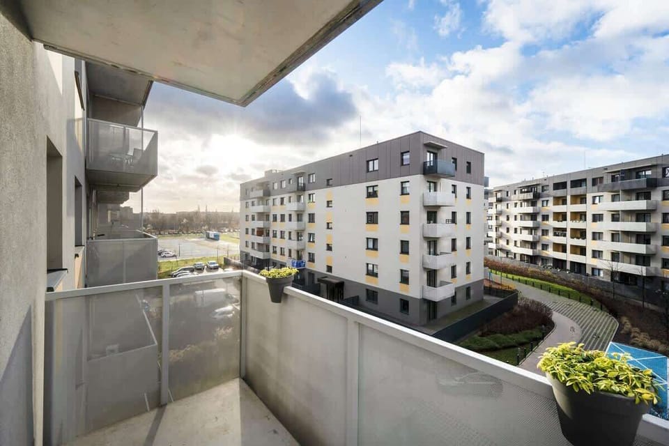 A balcony with grey concrete railings overlooking a residential complex with landscaped gardens and neighboring apartment buildings.
