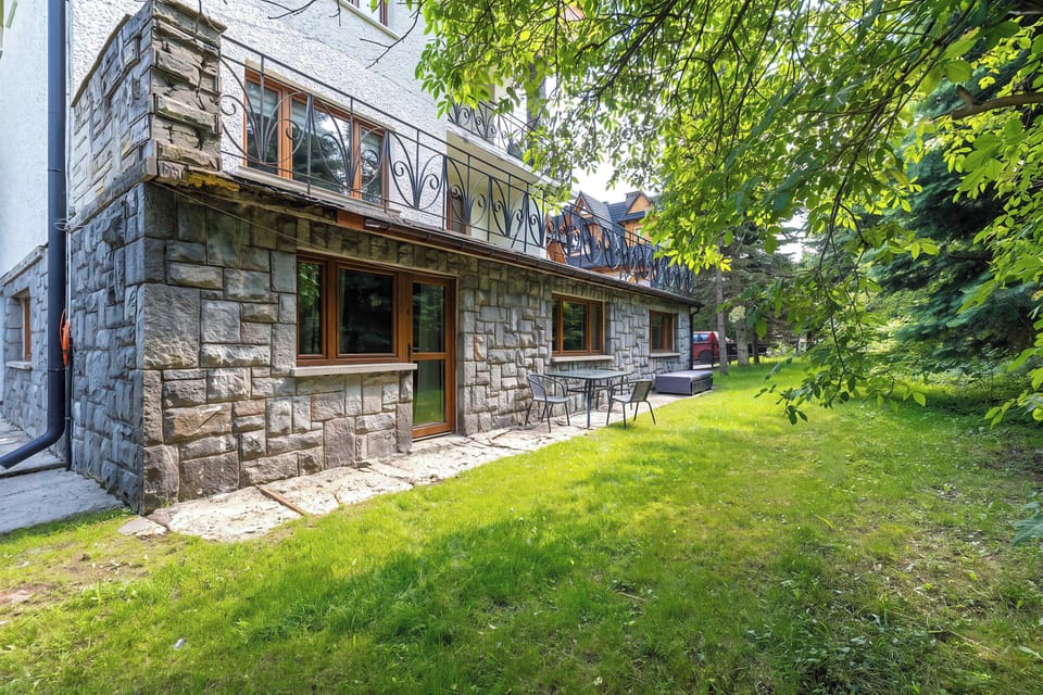 A wide angle showcases the rustic stone architecture of the house facing a sprawling green garden. Tall trees provide shade over the lawn.