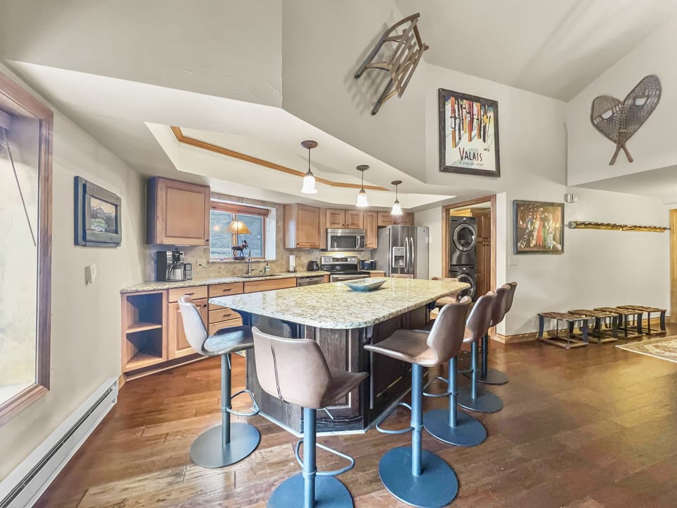 A spacious kitchen with wooden cabinetry, a large granite island with brown barstools, stainless steel appliances, and various wall decorations, including framed art and snowshoes.
