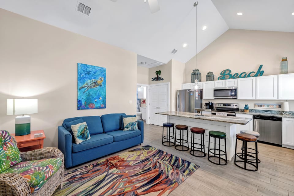 Relax in your bright, coastal-themed living space with vibrant blue sofa and open kitchen featuring colorful bar stools for memorable gatherings.